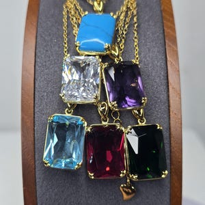 May include: A collection of gold-tone necklaces featuring rectangular pendants with various coloured stones, including turquoise, clear, purple, light blue, deep red, and dark green, all suspended from delicate gold-tone metal chains.