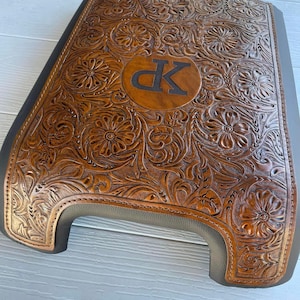 May include: A brown leather vehicle center console lid cover featuring an intricate embossed floral pattern and a central circular emblem containing the letters XP. The cover has dark brown stitched edges and a smooth dark gray border.