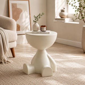 May include: A cream-coloured side table with a unique human-like form. The table has a round top and a base shaped like a person sitting with their legs extended. A small vase, a candle, and a book with the text "LIVE BEAUTIFUL" are on top.