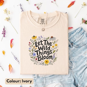 May include: An ivory t-shirt featuring a graphic print of colorful wildflowers surrounding the black text Let The Wild Things Bloom. The shirt has a crew neckline and a Comfort Colors brand label at the inner collar, paired with blue denim shorts.