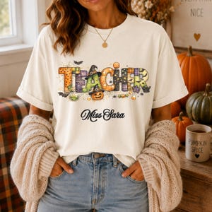 Puede incluir: Una camiseta de manga corta de color crema con la palabra TEACHER en letras de bloque coloridas y estampadas, decoradas con motivos de Halloween como calabazas, murciélagos y telarañas. Debajo, el texto Miss Sara está impreso en cursiva negra.
