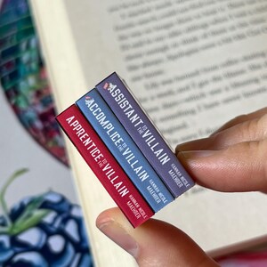 May include: A stack of three miniature books with the titles "Apprentice to the Villain", "Accomplice to the Villain", and "Assistant to the Villain". The books are red, blue, and purple, respectively, and held near an open book.