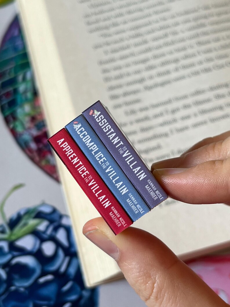 May include: A stack of three miniature books with the titles "Apprentice to the Villain", "Accomplice to the Villain", and "Assistant to the Villain". The books are red, blue, and purple, respectively, and held near an open book.