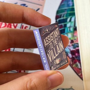 May include: A miniature book titled "Assistant to the Villain" held between fingers. The book has a purple cover with the title in white and a cityscape illustration. The author's name, Hannah Nicole Maehrer, is also visible.