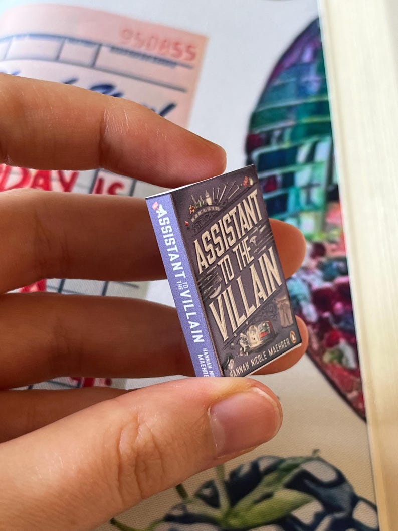 May include: A miniature book titled "Assistant to the Villain" held between fingers. The book has a purple cover with the title in white and a cityscape illustration. The author's name, Hannah Nicole Maehrer, is also visible.