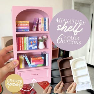Könnte beinhalten: Ein rosa Miniatur-Bücherregal mit einer Schublade, gefüllt mit winzigen Büchern und einer kleinen Vase mit Blumen. Der Text "MINIATURE shelf 6 COLOR OPTIONS" befindet sich in einem lila Kreis. Die Schublade öffnet sich.