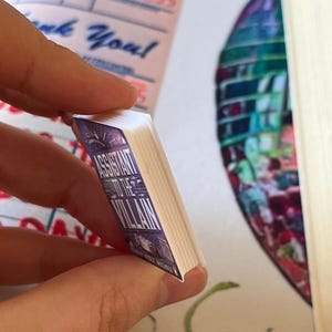 May include: A miniature book titled "Assistant to the Villain" with a purple and white cover. The book is held between fingers, showcasing its small size and detailed design. The book's pages are white.