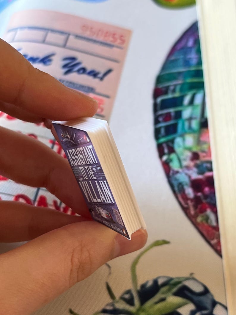 May include: A miniature book titled "Assistant to the Villain" with a purple and white cover. The book is held between fingers, showcasing its small size and detailed design. The book's pages are white.