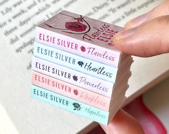 May include: A stack of miniature books with pastel-colored spines. Each book features the author's name, "ELSIE SILVER," and a title: "Flawless," "Heartless," "Powerless," "Reckless," and "Hopeless." The books are held in front of an open book.