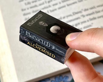 May include: Two miniature books are stacked, held against an open book. The top book is titled "MANACLED" with an illustration of a person. The bottom book is titled "ALCHEMISED". The books are black with gold lettering.