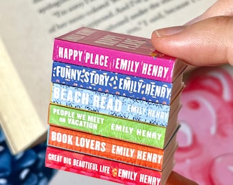 May include: A stack of miniature books with colorful spines, held by a hand. The titles include "HAPPY PLACE," "FUNNY STORY," "BEACH READ," "PEOPLE WE MEET ON VACATION," "BOOK LOVERS," and "GREAT BIG BEAUTIFUL LIFE" by Emily Henry.