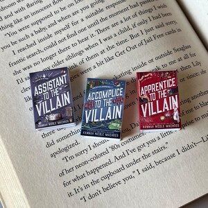 May include: Three miniature books titled "Assistant to the Villain," "Accomplice to the Villain," and "Apprentice to the Villain" are displayed open on a book page. The book covers are colorful with text and illustrations. The author's name, Hannah Nicole Maehrer, is printed on each book.
