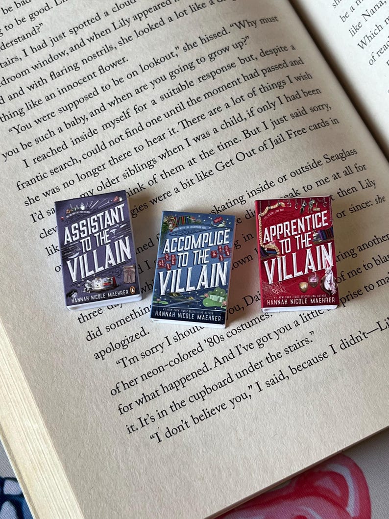 May include: Three miniature books titled "Assistant to the Villain," "Accomplice to the Villain," and "Apprentice to the Villain" are displayed open on a book page. The book covers are colorful with text and illustrations. The author's name, Hannah Nicole Maehrer, is printed on each book.
