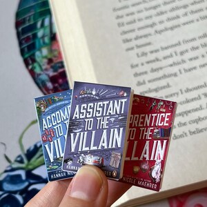 May include: Three miniature books with colorful covers are held in front of an open book. The titles are "Accompany to the Villain", "Assistant to the Villain", and "Prentice to the Villain". The books have detailed illustrations and text.