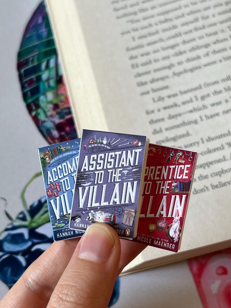 May include: Three miniature books with colorful covers are held in front of an open book. The titles are "Accompany to the Villain", "Assistant to the Villain", and "Prentice to the Villain". The books have detailed illustrations and text.