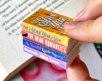 May include: A stack of four miniature books with colorful covers. The top book is titled "The Seven Year" in yellow and red. Below are "The Seven Year Slip", "The Dead Romantics", "A Novel Love Story", and "Sounds Like Love".
