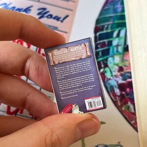 May include: A miniature book with the title "ASSISTANT WANTED" on the cover. The back of the book is visible, with text and a small illustration of a frog. The book is held in a person's hand, with a "Thank You" receipt in the background.