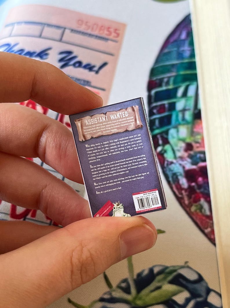 May include: A miniature book with the title "ASSISTANT WANTED" on the cover. The back of the book is visible, with text and a small illustration of a frog. The book is held in a person's hand, with a "Thank You" receipt in the background.