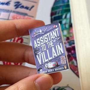 May include: A miniature book titled "Assistant to the Villain" with a purple cover. The title is in white, with the author's name, Hannah Nicole Maehrer, at the bottom. The book has a Penguin logo and illustrations.