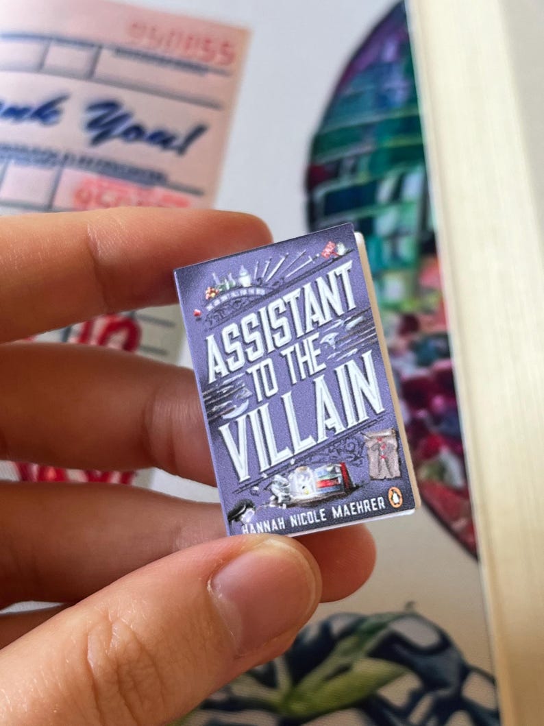 May include: A miniature book titled "Assistant to the Villain" with a purple cover. The title is in white, with the author's name, Hannah Nicole Maehrer, at the bottom. The book has a Penguin logo and illustrations.