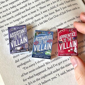 May include: Three miniature book replicas with the titles "Assistant to the Villain," "Accomplice to the Villain," and "Apprentice to the Villain." The books have colorful covers and are held over an open book with printed text.