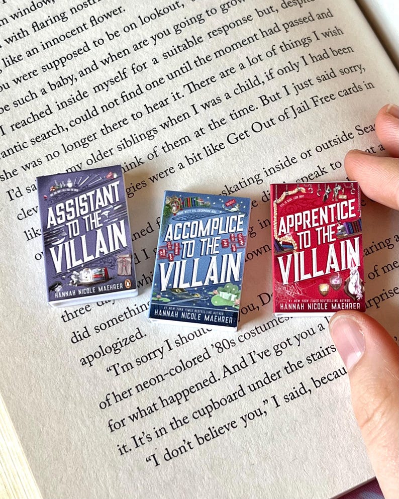 May include: Three miniature book replicas with the titles "Assistant to the Villain," "Accomplice to the Villain," and "Apprentice to the Villain." The books have colorful covers and are held over an open book with printed text.