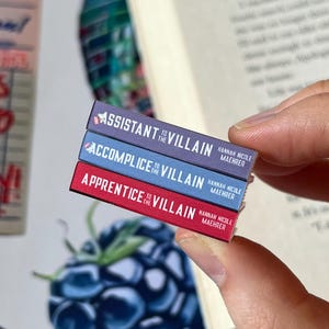 May include: A stack of three miniature books with the titles "Assistant to the Villain", "Accomplice to the Villain", and "Apprentice to the Villain". Each book has a different colored spine: purple, blue, and red. The author's name, Hannah Nicole Maehrer, is printed on each book.