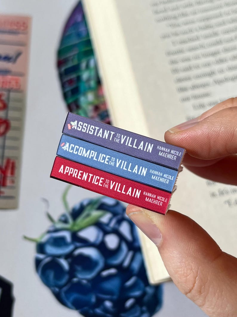 May include: A stack of three miniature books with the titles "Assistant to the Villain", "Accomplice to the Villain", and "Apprentice to the Villain". Each book has a different colored spine: purple, blue, and red. The author's name, Hannah Nicole Maehrer, is printed on each book.