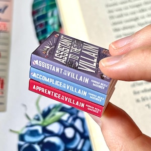May include: A stack of three miniature books with the titles "Assistant to the Villain", "Accomplice to the Villain", and "Apprentice to the Villain". The books are in purple, blue, and red, with white text. The author's name, Hannah Nicole Maehrer, is also visible.