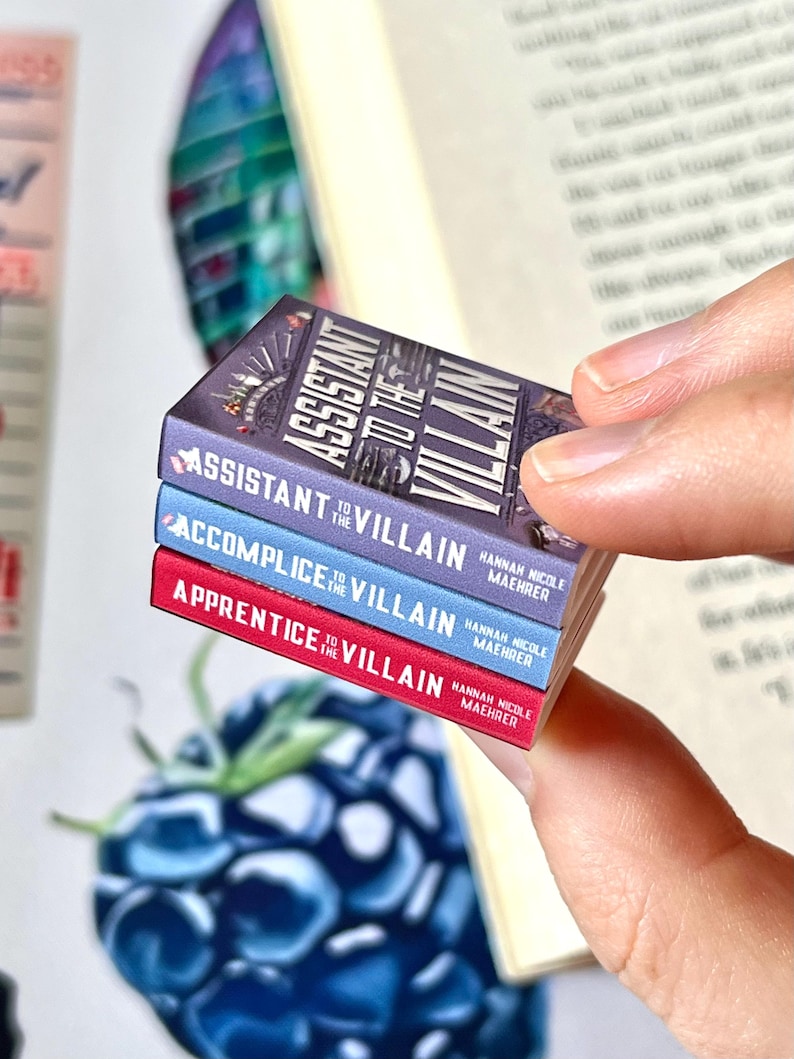 May include: A stack of three miniature books with the titles "Assistant to the Villain", "Accomplice to the Villain", and "Apprentice to the Villain". The books are in purple, blue, and red, with white text. The author's name, Hannah Nicole Maehrer, is also visible.