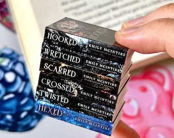 May include: A stack of miniature books with titles including "Hooked," "Wretched," and "Hexed." The books are black with white lettering and feature the author's name, Emily McIntire. The books are held in a person's hand.