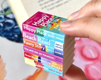 May include: A stack of miniature books with colorful covers, held in a hand. The titles include "Happy Place," "Beach Read," and "Funny Story" by Emily Henry. The books are stacked on top of each other.
