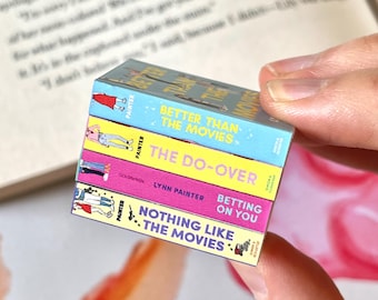 May include: A stack of four miniature books with colorful covers. The titles include "Better Than The Movies", "The Do-Over", "Betting on You", and "Nothing Like The Movies". The books are held in a person's hand, with a blurred book and floral background.