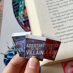 May include: Three miniature book replicas with titles including "Assistant to the Villain" and "Prentice to the Villain" are held in front of an open book. The book covers are colorful, with text and illustrations. The background includes a watercolor painting.