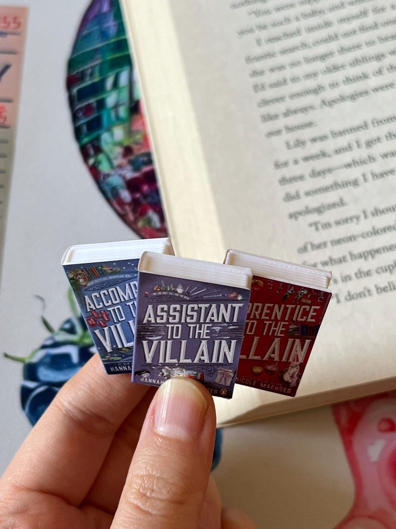 May include: Three miniature book replicas with titles including "Assistant to the Villain" and "Prentice to the Villain" are held in front of an open book. The book covers are colorful, with text and illustrations. The background includes a watercolor painting.