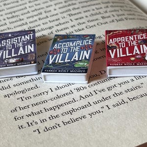 May include: Three miniature books with the titles "Assistant to the Villain," "Accomplice to the Villain," and "Apprentice to the Villain" are displayed on an open book. The book covers are colorful, with text and illustrations. The books are by Hannah Nicole Maehrer.