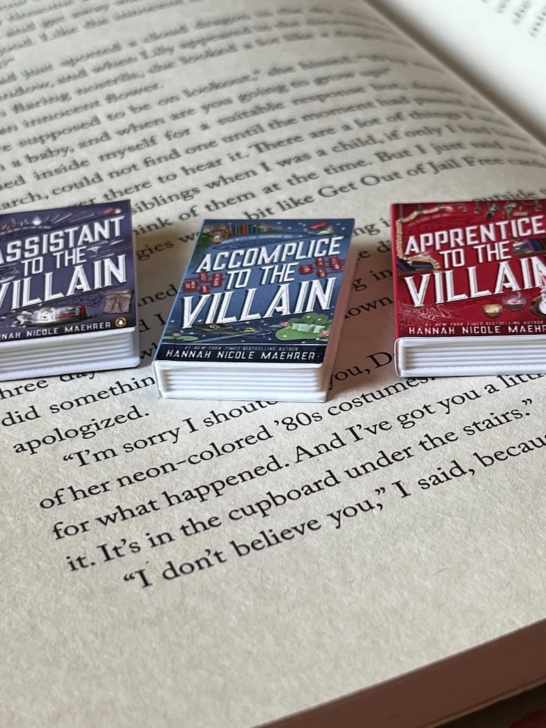 May include: Three miniature books with the titles "Assistant to the Villain," "Accomplice to the Villain," and "Apprentice to the Villain" are displayed on an open book. The book covers are colorful, with text and illustrations. The books are by Hannah Nicole Maehrer.