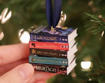 May include: A miniature stack of five books hanging from a dark blue ribbon. The book spines are labeled Eragon, Eldest, Brisingr, Inheritance, and Murtagh. Each book has a distinct color spine including blue, red, brown, teal, and black.