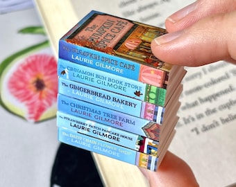 May include: A stack of miniature books with colorful covers. The titles include "The Pumpkin Spice Cafe," "Cinnamon Bun Book Store," and "The Gingerbread Bakery." Each book is held in a hand, with a larger book in the background.