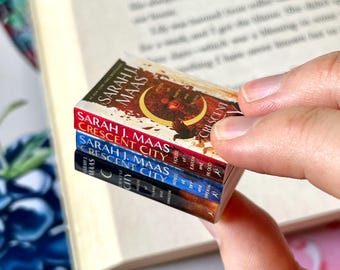 May include: A stack of three miniature books with the titles "Crescent City" by Sarah J. Maas. The books have red, blue, and black spines. The covers feature illustrations and text. The books are held near an open book.