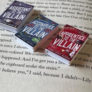 May include: Three miniature books with the titles "Assistant to the Villain," "Accomplice to the Villain," and "Apprentice to the Villain." The books are on a page of printed text. Each book has a different color cover.