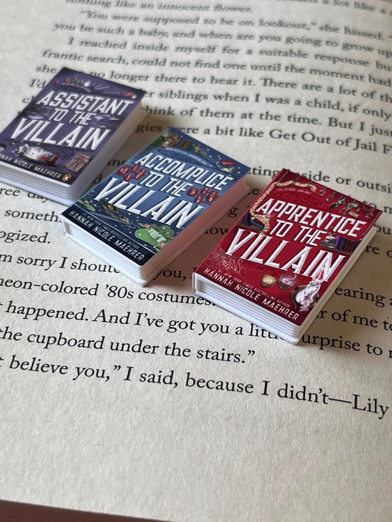 May include: Three miniature books with the titles "Assistant to the Villain," "Accomplice to the Villain," and "Apprentice to the Villain." The books are on a page of printed text. Each book has a different color cover.