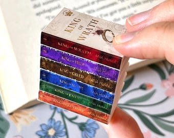 May include: A stack of miniature books with colorful spines. The top book reads "KING OF WRATH" with a gold ring illustration. The other books have titles like "KING OF PRIDE" and "KING OF GREED" with the author's name, Ana Huang.