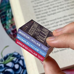May include: A stack of three miniature books with the titles "Assistant to the Villain," "Accomplice to the Villain," and "Apprentice to the Villain." The books have colorful spines and detailed cover art, held near an open book.