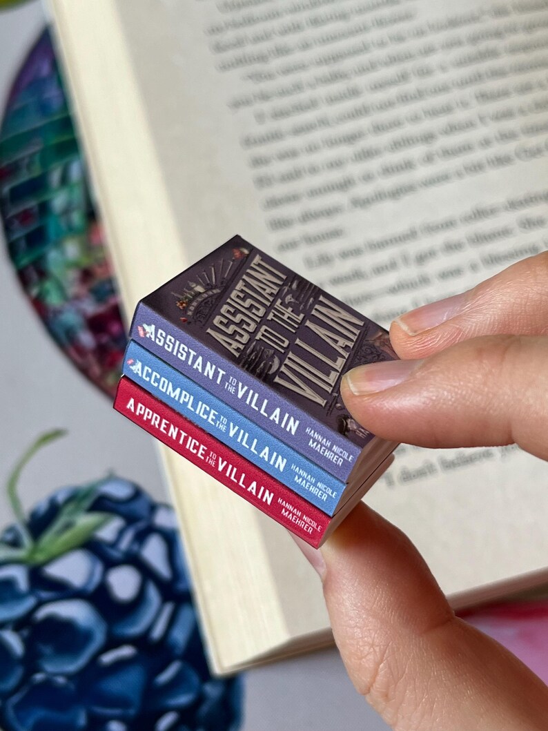 May include: A stack of three miniature books with the titles "Assistant to the Villain," "Accomplice to the Villain," and "Apprentice to the Villain." The books have colorful spines and detailed cover art, held near an open book.
