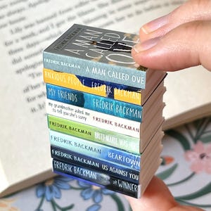 May include: A stack of miniature books held in a hand. The book titles include "A Man Called Ove", "Anxious People", "My Friends", "Britt-Marie Was Here", "Beartown", "Us Against You", and "The Winner". The author's name, Fredrik Backman, is visible on each spine.