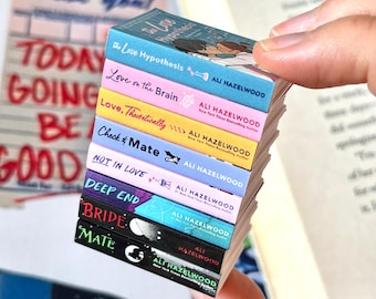 May include: A stack of miniature books with colorful covers, held in a hand. The titles include "The Love Hypothesis," "Love on the Brain," and "Love, Theoretically." The author's name, Ali Hazelwood, is printed on each book.