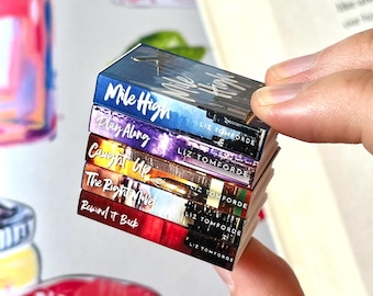 May include: A stack of miniature books with colorful covers. The titles include "Mile High," "Play Along," "Caught Up," "The Right Move," and "Rewind it Back." Each book has the author's name, "LIZ TOMFORDE," printed on the spine. The books are held in a person's hand.