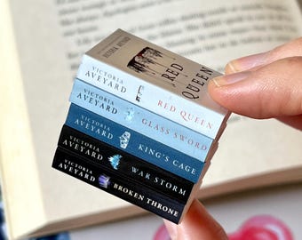 May include: A stack of miniature books from the "Red Queen" series by Victoria Aveyard. The titles include "Red Queen", "Glass Sword", "King's Cage", "War Storm", and "Broken Throne". The books are held in a person's hand, with an open book in the background.