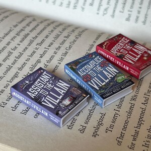 May include: Three miniature books with the titles "Assistant to the Villain," "Accomplice to the Villain," and "Apprentice to the Villain" are displayed on an open book. The books have colorful covers with illustrations and text. The pages of the open book are visible in the background.